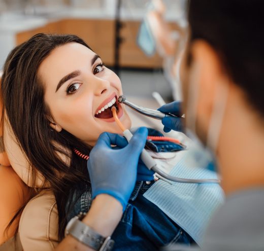 young-female-patient-visiting-dentist-office Young female patient visiting dentist office. Beautiful woman with healthy straight white teeth sitting at dental chair with open mouth during oral checkup while doctor working at teeth.